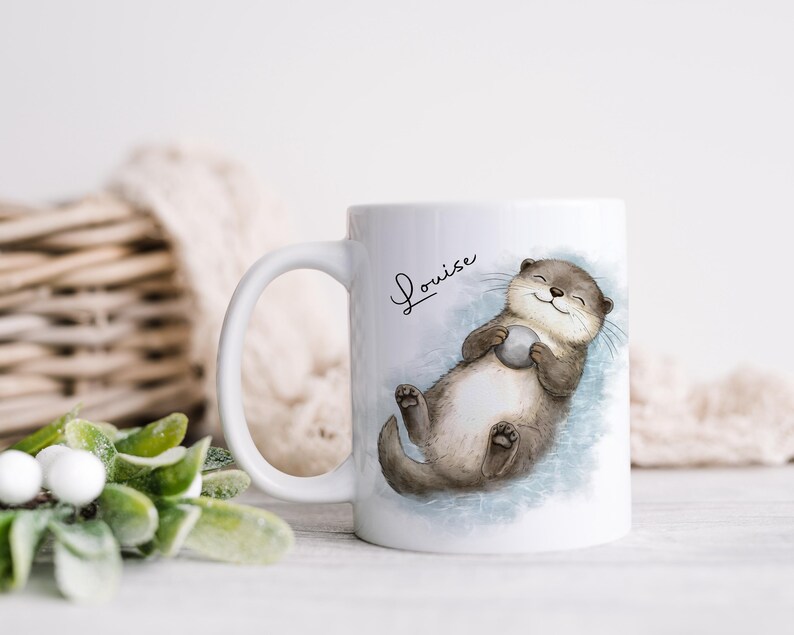 Pu&ograve; includere: Una tazza in ceramica bianca con l'illustrazione di una lontra marrone che galleggia sulla schiena tenendo una piccola pietra grigia. Il nome Louise &egrave; stampato in corsivo nero sopra la lontra, su uno sfondo ad acquerello blu tenue.