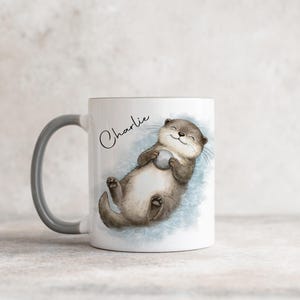 Pu&ograve; includere: Una tazza in ceramica bianca con manico grigio, caratterizzata da un'illustrazione ad acquerello di una lontra marrone che galleggia sulla schiena tenendo un piccolo sasso grigio. Il nome Charlie &egrave; scritto in corsivo nero sopra l'illustrazione.