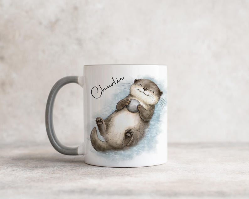 Pu&ograve; includere: Una tazza in ceramica bianca con manico grigio, caratterizzata da un'illustrazione ad acquerello di una lontra marrone che galleggia sulla schiena tenendo un piccolo sasso grigio. Il nome Charlie &egrave; scritto in corsivo nero sopra l'illustrazione.