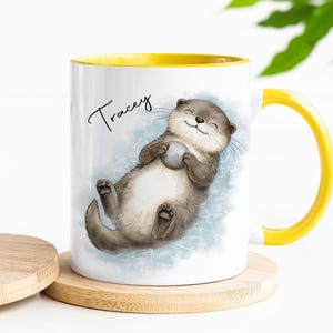Pu&ograve; includere: Una tazza in ceramica bianca con manico e bordo interno giallo brillante. L'esterno presenta un'illustrazione ad acquerello di una lontra marrone che galleggia sulla schiena tenendo una piccola pietra grigia. Il nome Tracey &egrave; stampato in nero.