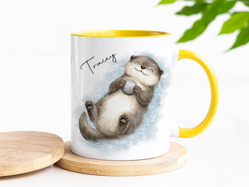 Pu&ograve; includere: Una tazza in ceramica bianca con manico e bordo interno giallo brillante. L'esterno presenta un'illustrazione ad acquerello di una lontra marrone che galleggia sulla schiena tenendo una piccola pietra grigia. Il nome Tracey &egrave; stampato in nero.