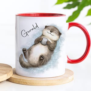 Pu&ograve; includere: Una tazza in ceramica bianca con interno e manico rosso brillante. L'esterno presenta un'illustrazione in stile acquerello di una lontra marrone che galleggia sulla schiena tenendo una piccola pietra grigia. Il nome Gerald &egrave; stampato in nero.