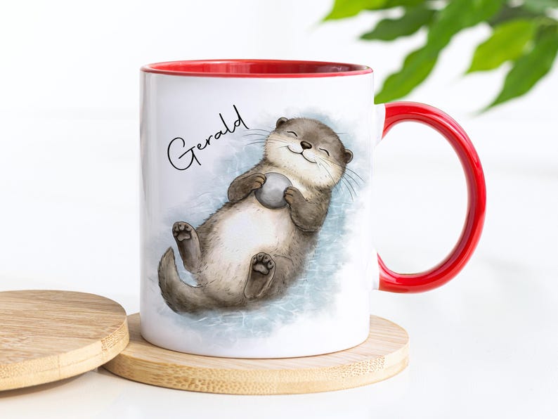 Pu&ograve; includere: Una tazza in ceramica bianca con interno e manico rosso brillante. L'esterno presenta un'illustrazione in stile acquerello di una lontra marrone che galleggia sulla schiena tenendo una piccola pietra grigia. Il nome Gerald &egrave; stampato in nero.