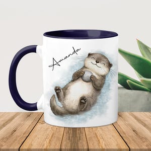 Pu&ograve; includere: Una tazza in ceramica bianca con interno e manico blu scuro, caratterizzata da un'illustrazione ad acquerello di una lontra marrone che galleggia sulla schiena tenendo una piccola pietra grigia, con il nome Amanda scritto in nero sopra.