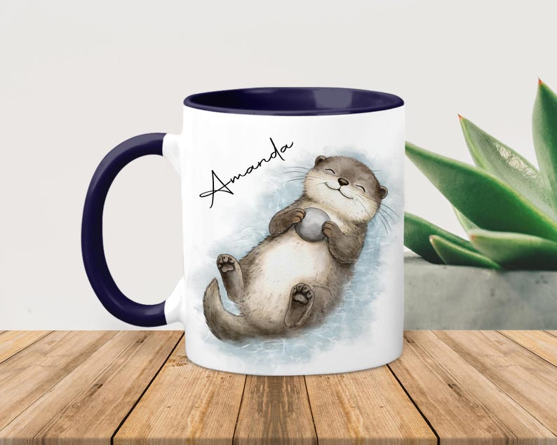 Pu&ograve; includere: Una tazza in ceramica bianca con interno e manico blu scuro, caratterizzata da un'illustrazione ad acquerello di una lontra marrone che galleggia sulla schiena tenendo una piccola pietra grigia, con il nome Amanda scritto in nero sopra.
