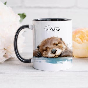 May include: A white ceramic mug with a black handle and interior rim. The mug features a printed illustration of a brown otter sleeping on a blue surface. The name Peter is printed in black cursive script above the otter illustration.
