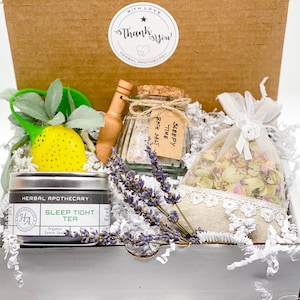 May include: A gift box filled with relaxation items, including a jar of bath salts labeled "Sleepy Time Bath Salt", a bag of dried herbs, a tea tin labeled "Sleep Tight Tea", and a small wooden spoon.