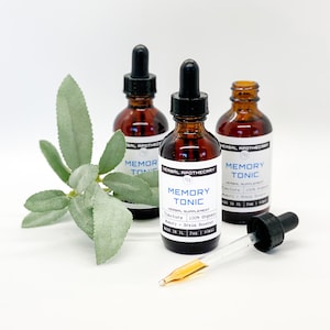 May include: Three brown glass bottles with droppers filled with a clear liquid. The bottles are labeled "Memory Tonic" and "Herbal Supplement 100% Organic" with a blue and white label. The bottles are sitting on a white surface with a green leafy plant in the background.