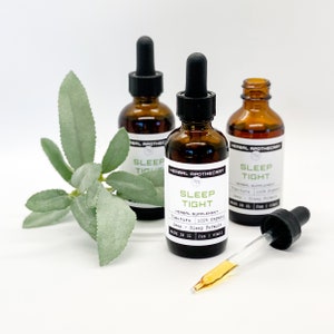 May include: Three amber glass bottles with droppers, labeled "Sleep Tight" and "Herbal Apothecary". The bottles are on a white surface with a sprig of green foliage.