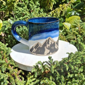 Könnte beinhalten: Eine Keramik-Tasse mit Bergmotiv. Die Tasse hat einen blau-weißen Farbverlauf mit einer Bergketten-Illustration in Schwarz und Beige. Der Griff ist blau und die Tasse steht auf einer weißen, runden Oberfläche.