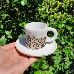 May include: White ceramic coffee cup and saucer with a black and white mushroom design. The cup has a handle and the saucer is round.