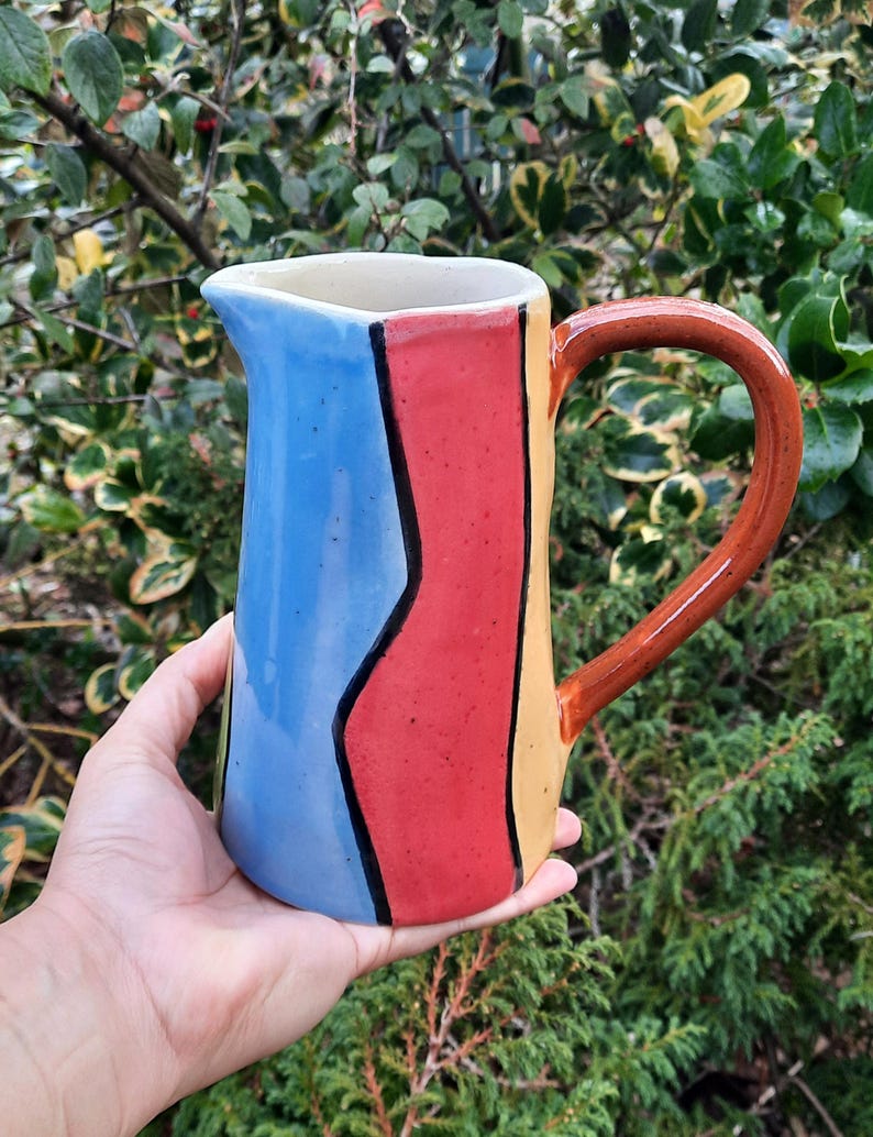 Op de afbeelding: Een kleurrijke keramische kan met een blauw, rood en geel gestreept ontwerp. De kan heeft een bruin handvat en een witte rand.