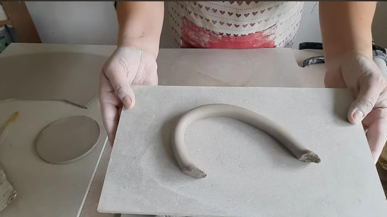 Op de afbeelding: Een close-up van een grijze keramische handgreep, gevormd als een gebogen boog, liggend op een wit betegeld oppervlak.