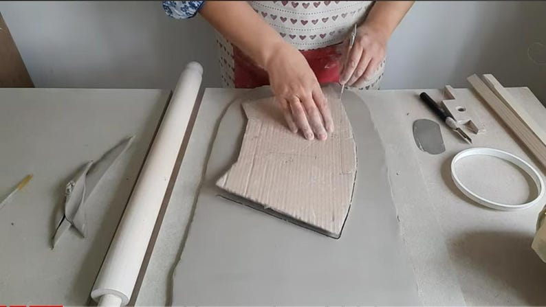 Op de afbeelding: Een persoon gebruikt een gereedschap om een ​​stuk klei te snijden. De klei ligt op een tafel met een deegroller, een stuk klei en een ring.