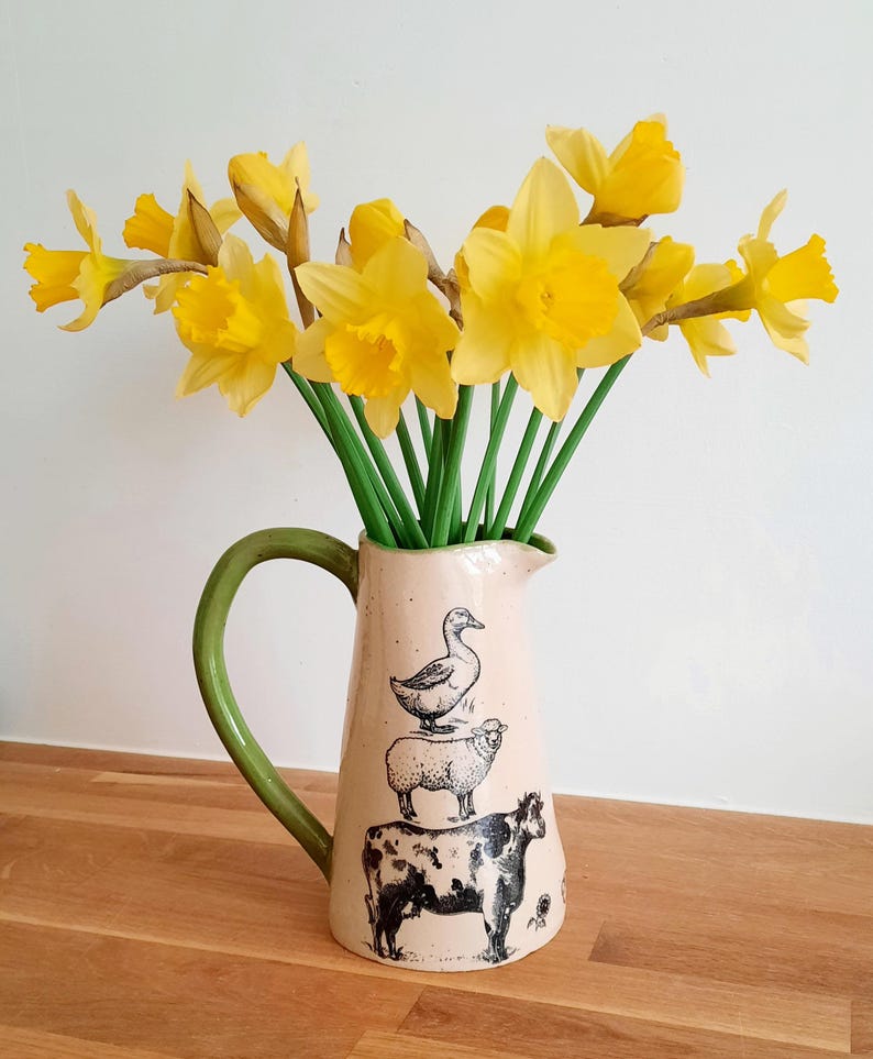Op de afbeelding: Een keramische kan met een groene handvat en een zwart-witte illustratie van een koe, een schaap en een eend. De kan is gevuld met een boeket gele narcissen.