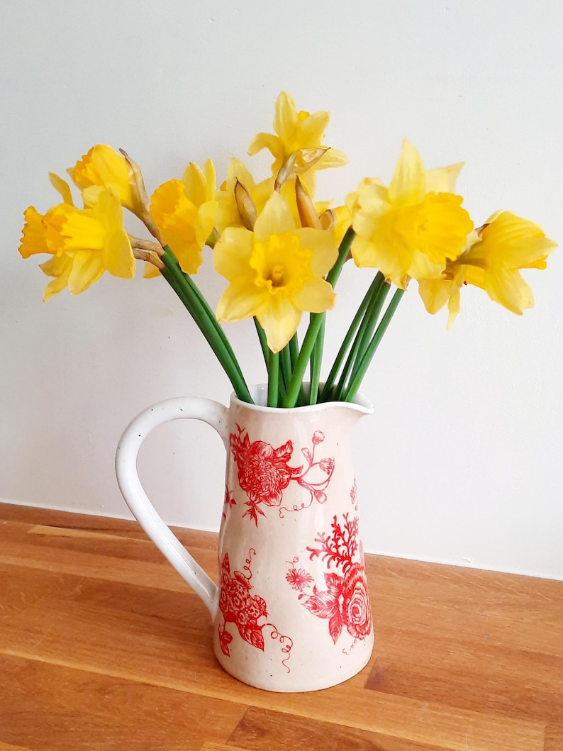 Op de afbeelding: Een witte keramische kan met een rood bloemenpatroon, gevuld met een bos gele narcissen.