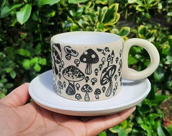 May include: A white ceramic mug with a black mushroom design. The mug has a handle and is sitting on a white saucer.