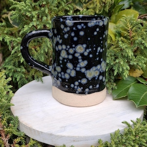 May include: A ceramic mug with a black speckled glaze and a natural beige base. The mug has a large, curved handle. It is approximately 10 cm tall. The speckled pattern includes blue and tan spots, creating a unique, artistic design.