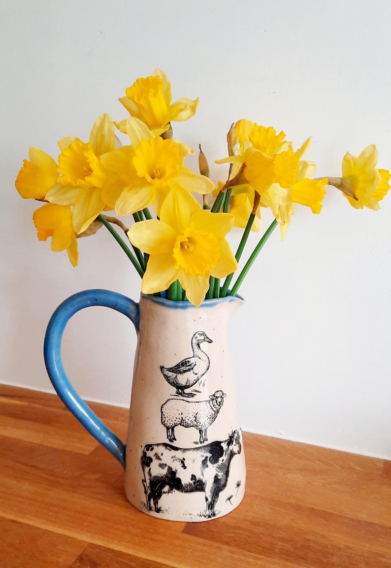 Op de afbeelding: Een blauwe en witte keramische kan met een eend, schaap en koe ontwerp. De kan is gevuld met een boeket gele narcissen.