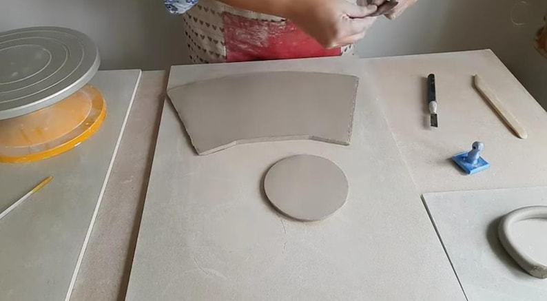 Op de afbeelding: Een pottenbakkersstudio met een pottenbakkerswiel, een keramische schijf, een keramische kom en een keramisch gereedschap. De gereedschappen bevinden zich op een wit oppervlak.