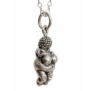 Puede incluir: Un collar con colgante plateado que presenta una peque&ntilde;a figura estilizada de una mujer. La figura tiene una forma redondeada con rasgos exagerados y est&aacute; suspendida de una delicada cadena.