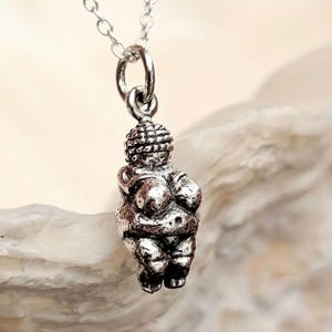 A silver pendant necklace with a miniature sculpture of the Venus of Willendorf. The pendant, suspended from a fine silver chain, depicts a stylised, curvaceous female form. The sculpture is highly detailed.