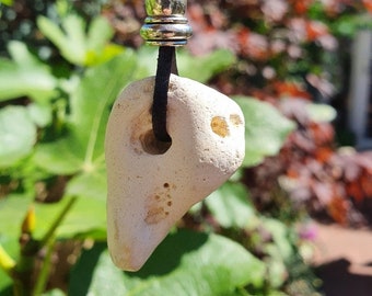 May include: A pendant necklace featuring a cream-colored, naturally shaped stone with a hole. The stone is suspended from a black cord, accented with a silver-toned bead. The background is blurred with green and red foliage.