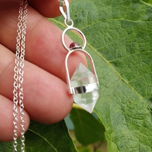 Op de afbeelding: Een zilveren ketting met een hanger van een helder kristalpunt en een kleine rode edelsteen. Het kristalpunt hangt in een zilveren kooi.