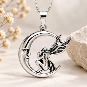 May include: A silver-toned metal pendant necklace featuring a crescent moon with a human-like face and a winged fairy figure sitting inside the curve of the moon. The pendant hangs from a delicate silver-toned chain with a polished finish.
