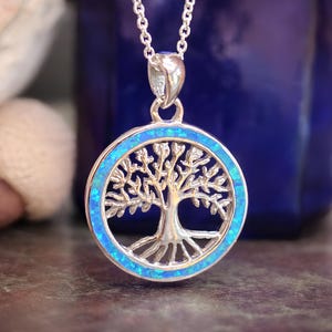 May include: A silver-toned circular pendant necklace featuring a tree of life design. The outer ring of the pendant is inlaid with bright blue iridescent opal-like material. The pendant hangs from a delicate silver-toned chain link necklace.