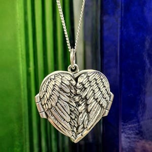 May include: A silver heart-shaped locket pendant suspended from a delicate silver chain. The front of the locket features a detailed, raised relief design of two symmetrical angel wings that meet in the centre, creating a textured metallic surface.