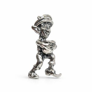 Op de afbeelding: Een zilveren bedel met een glimlachende kabouter. De figuur draagt een hoed, jas, broek en schoenen. De kabouter houdt een pot of bundel vast. De bedel heeft een gedetailleerd, vintage uiterlijk.