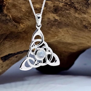 May include: A silver pendant necklace with a Celtic knot design. The pendant has a central, round, iridescent stone. The necklace chain is also silver. The pendant is set against a blurred wooden background.