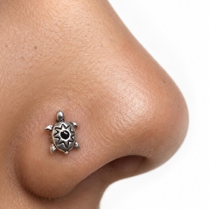 May include: A silver-toned nose stud featuring a turtle-shaped design. The turtle has a polished metallic finish with a central black circular inlay surrounded by a starburst pattern on its shell, creating a detailed and textured appearance.