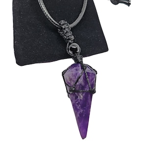 May include: A purple amethyst crystal pendant necklace. The crystal is encased in a black woven net and hangs from a black cord. A black bead is positioned above the crystal. The necklace is displayed against a black pouch.