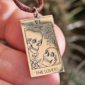 May include: Gold-colored rectangular pendant necklace featuring a tarot card design. The card depicts two skulls, floral accents, and the text "THE LOVERS" at the bottom. The pendant is suspended from a brown cord.