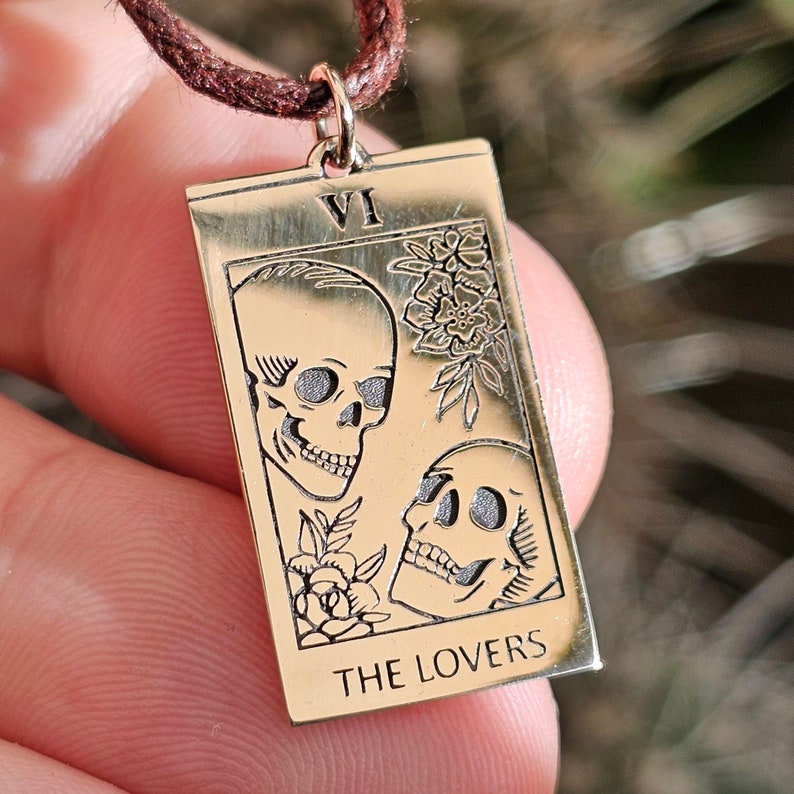 May include: Gold-colored rectangular pendant necklace featuring a tarot card design. The card depicts two skulls, floral accents, and the text "THE LOVERS" at the bottom. The pendant is suspended from a brown cord.