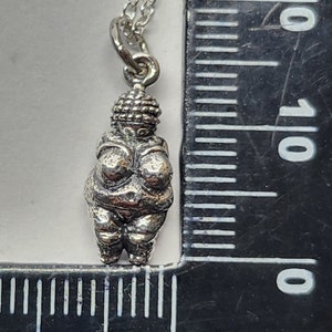 Puede incluir: Un collar con colgante de plata con una peque&ntilde;a y detallada figura de la Venus de Willendorf. El colgante est&aacute; suspendido de una delicada cadena. El collar se muestra junto a una regla para la escala.