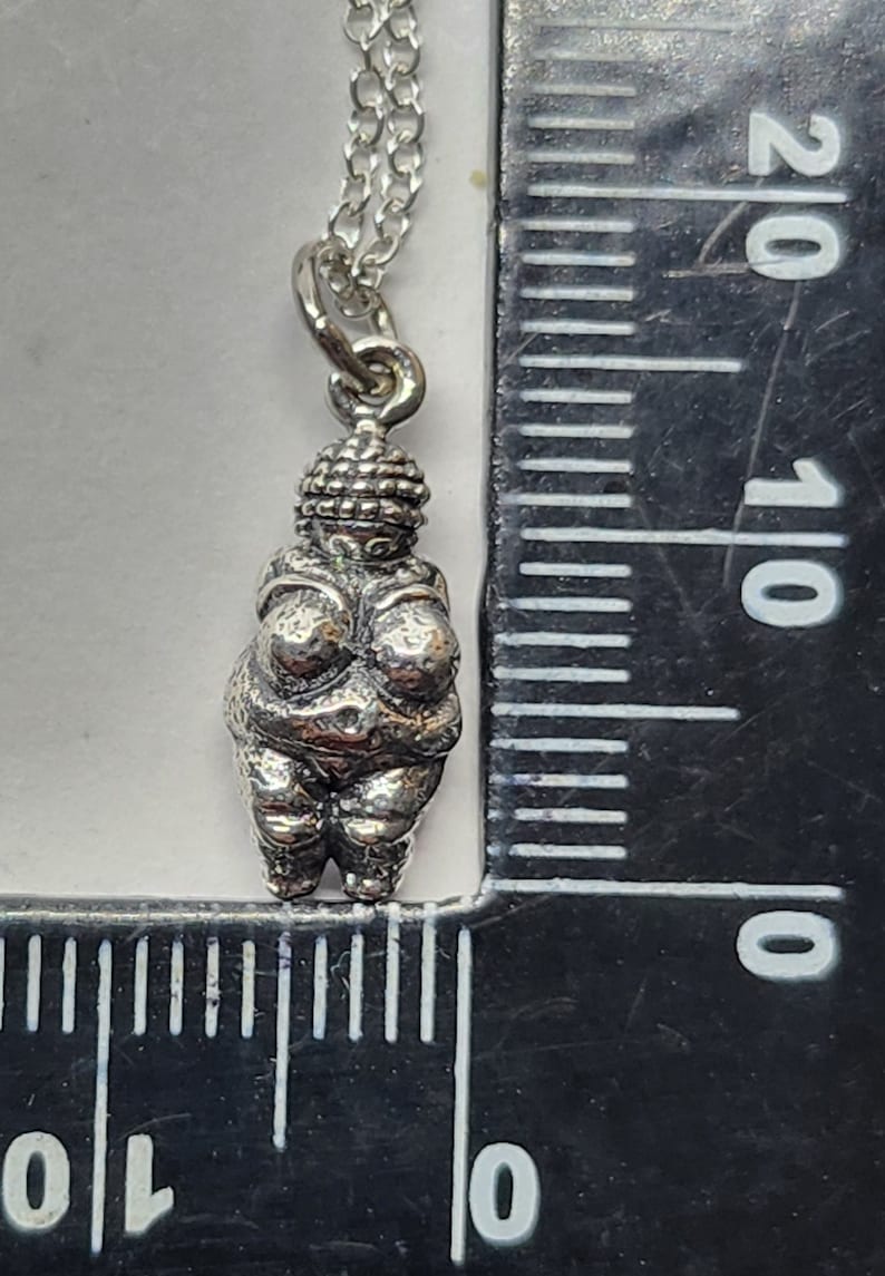 Puede incluir: Un collar con colgante de plata con una peque&ntilde;a y detallada figura de la Venus de Willendorf. El colgante est&aacute; suspendido de una delicada cadena. El collar se muestra junto a una regla para la escala.