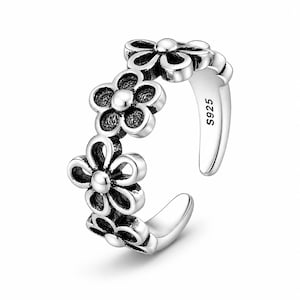 Può includere: Un anello aperto in argento caratterizzato da una serie di motivi floreali collegati con petali lucidi e centri scuri e testurizzati. La fascia interna è chiaramente stampata con il testo S925, che indica la composizione del materiale del gioiello.