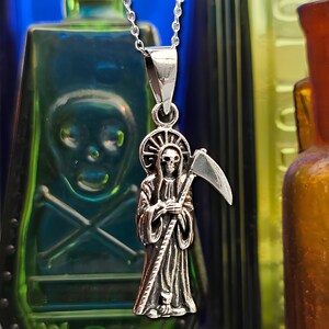 Pu&ograve; includere: Una collana con un ciondolo d'argento che raffigura una rappresentazione dettagliata della Morte che tiene una falce. Il ciondolo &egrave; appeso su uno sfondo di bottiglie di vetro colorate.