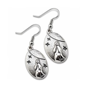 May include: A pair of silver-toned dangle earrings. Each earring features an oval pendant with a textured surface, a kneeling figure, and star accents. The earrings have a hook closure.