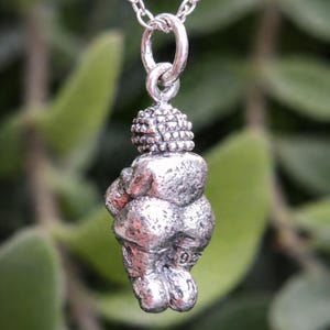 Puede incluir: Un collar con colgante de plata con una figura estilizada de una mujer. El colgante tiene una superficie texturizada y cuelga de una delicada cadena de plata. El fondo es un follaje verde borroso.