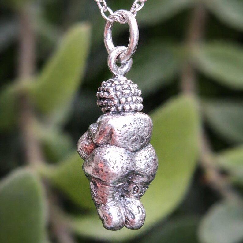 Puede incluir: Un collar con colgante de plata con una figura estilizada de una mujer. El colgante tiene una superficie texturizada y cuelga de una delicada cadena de plata. El fondo es un follaje verde borroso.