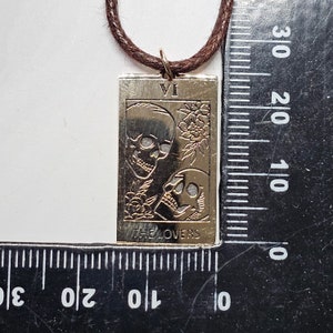 May include: A gold-colored rectangular pendant with the image of two skulls and floral accents, suspended from a brown cord. The pendant features the Roman numeral VI and the words "THE LOVERS". The pendant measures approximately 1.5 inches in length.
