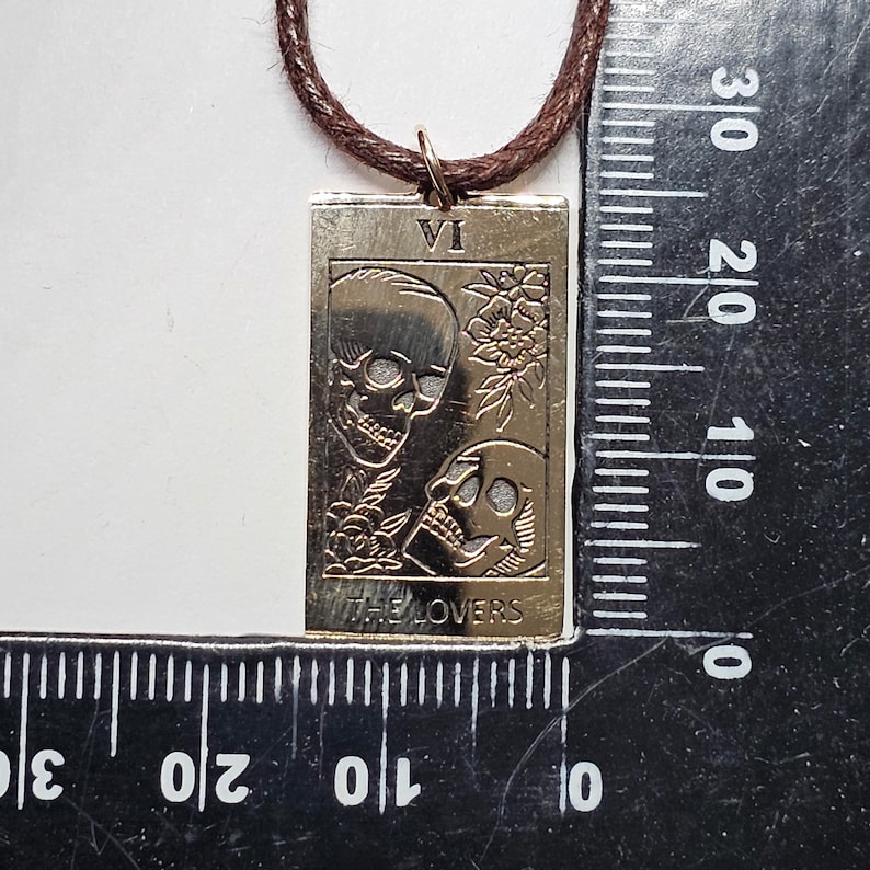 May include: A gold-colored rectangular pendant with the image of two skulls and floral accents, suspended from a brown cord. The pendant features the Roman numeral VI and the words "THE LOVERS". The pendant measures approximately 1.5 inches in length.