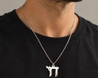 May include: A silver-toned metal pendant shaped like the Hebrew letter Chai, suspended from a matching thin silver-toned metal curb chain. The pendant features a polished, reflective surface and is worn against a plain black crew neck t-shirt.