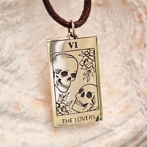 May include: A rectangular pendant necklace with a brown cord. The pendant is gold-colored and features an engraved tarot card design with two skulls, flowers, and the words "THE LOVERS" at the bottom. The Roman numeral "VI" is at the top.