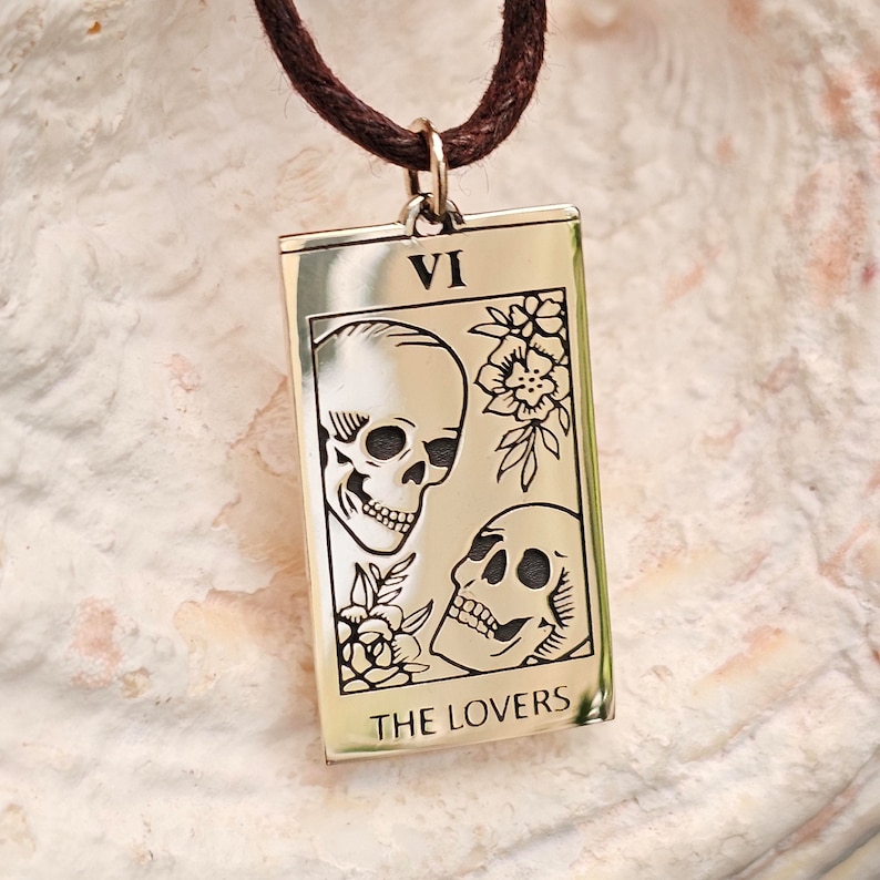May include: A rectangular pendant necklace with a brown cord. The pendant is gold-colored and features an engraved tarot card design with two skulls, flowers, and the words "THE LOVERS" at the bottom. The Roman numeral "VI" is at the top.