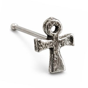 May include: A silver-toned nose stud featuring an ankh symbol with a textured, weathered surface. The jewellery piece has a straight post ending in a small spherical ball, designed for body piercing with a distinct Egyptian cross shape.